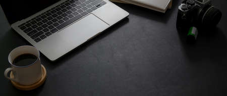 Cropped shot of dark modern office desk with laptop coffee cup camera and copy space on black table