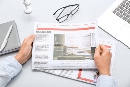 Woman reading newspaper at workplace with laptop and eyeglasses on gray background