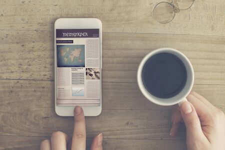 Reading newspaper from mobile phone holding coffee cup